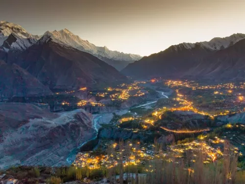 A wide valley surrounded by tall, snow-capped mountains is illuminated by clusters of warm lights from villages below at dusk. A river winds through the center of the valley, reflecting the fading light of the sky.